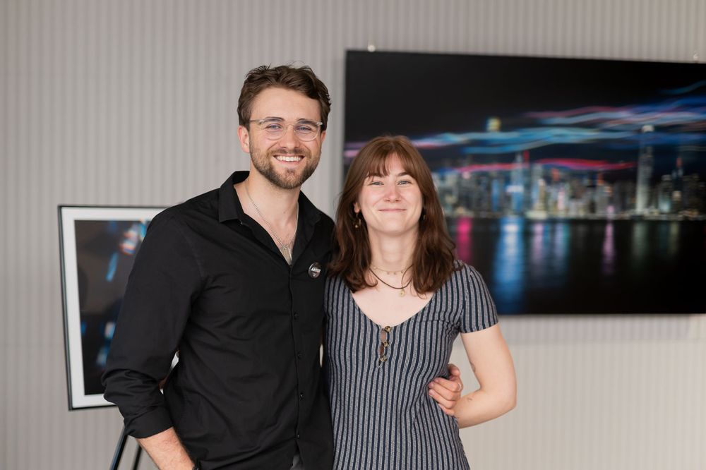 A smiling man and woman posing together in front of a colorful city skyline photograph at an indoor event.