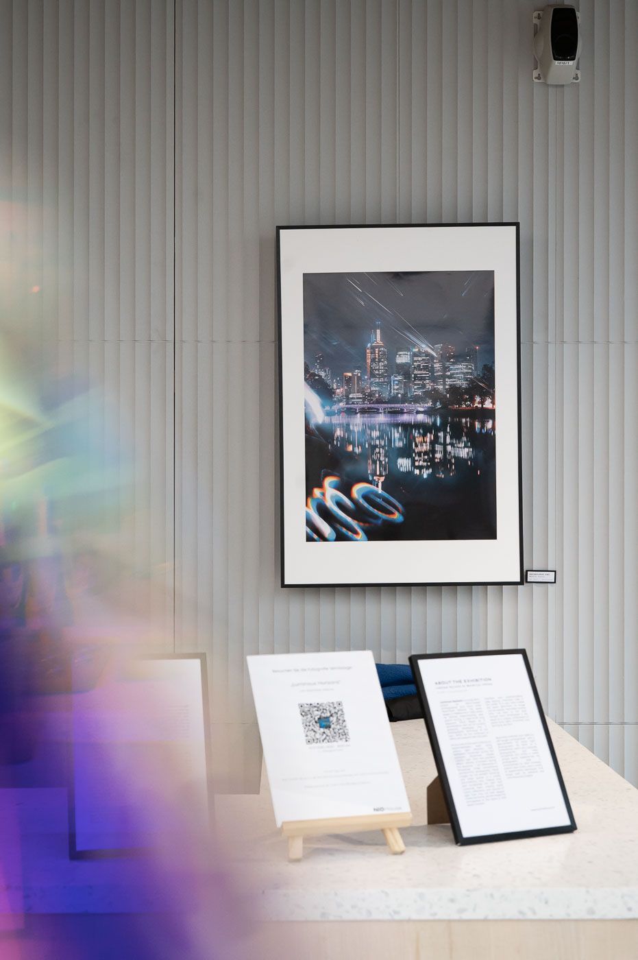 Framed cityscape night photography print displayed on a grey wall above informational placards at an art exhibition.