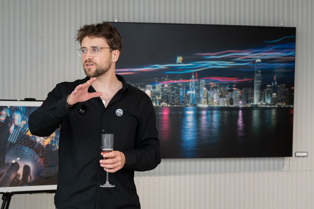 Man in black shirt holding champagne glass speaking at art gallery with cityscape photography exhibition.