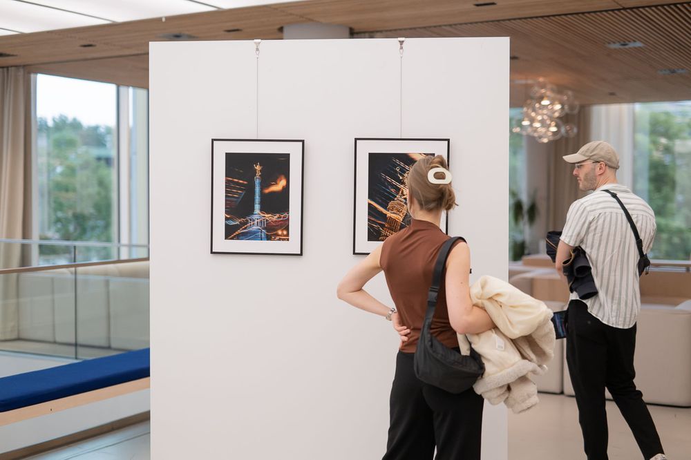 Two visitors viewing framed photography prints displayed on a white partition wall in a modern gallery space.