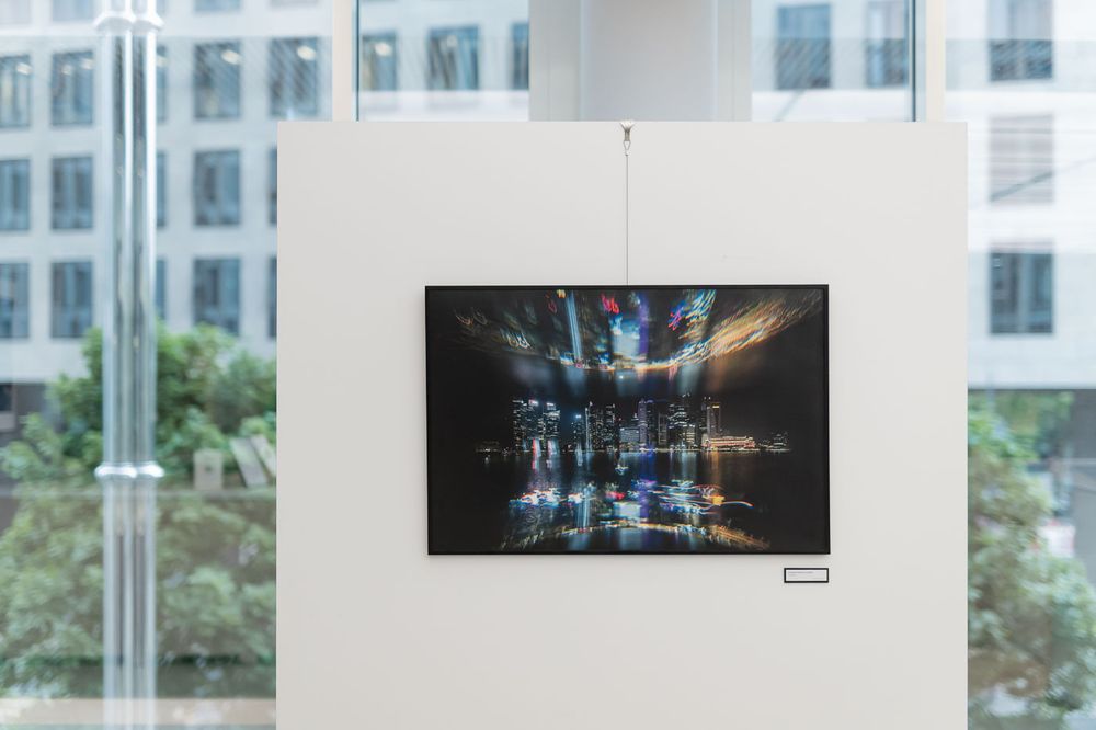 A framed night cityscape photograph displayed on a white gallery stand near large windows with green trees outside.