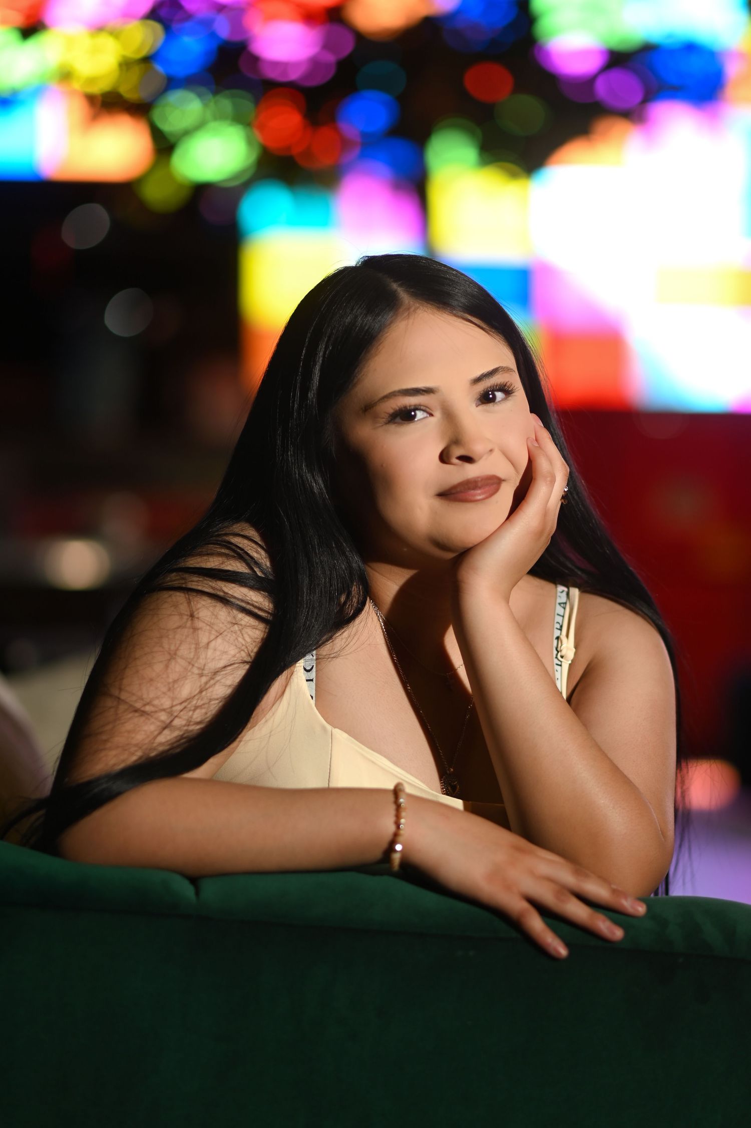 Young woman with long black hair rests her chin on her hand, posing against colorful bokeh lights background.