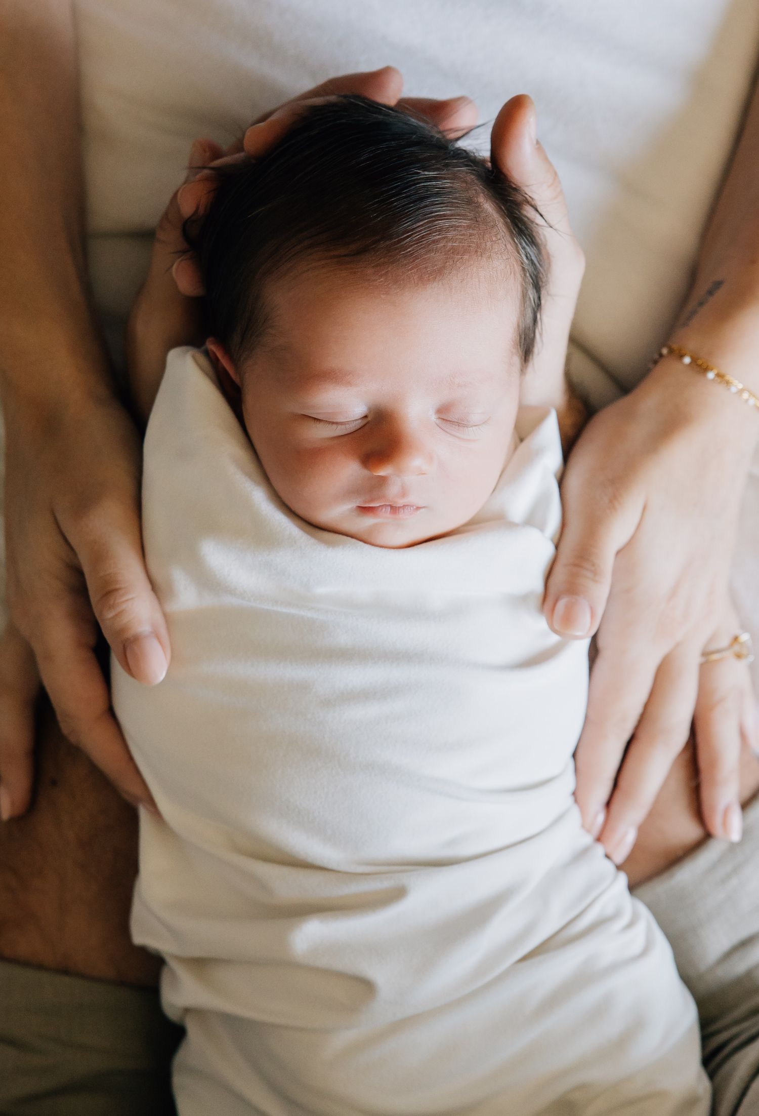 Newborn baby swaddled in white cloth sleeping peacefully while being cradled in parents hands.