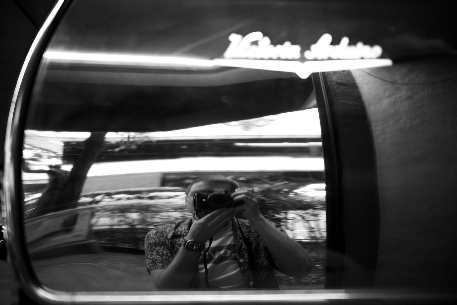 Black and white photo of a photographer's reflection in a vintage car mirror at night.