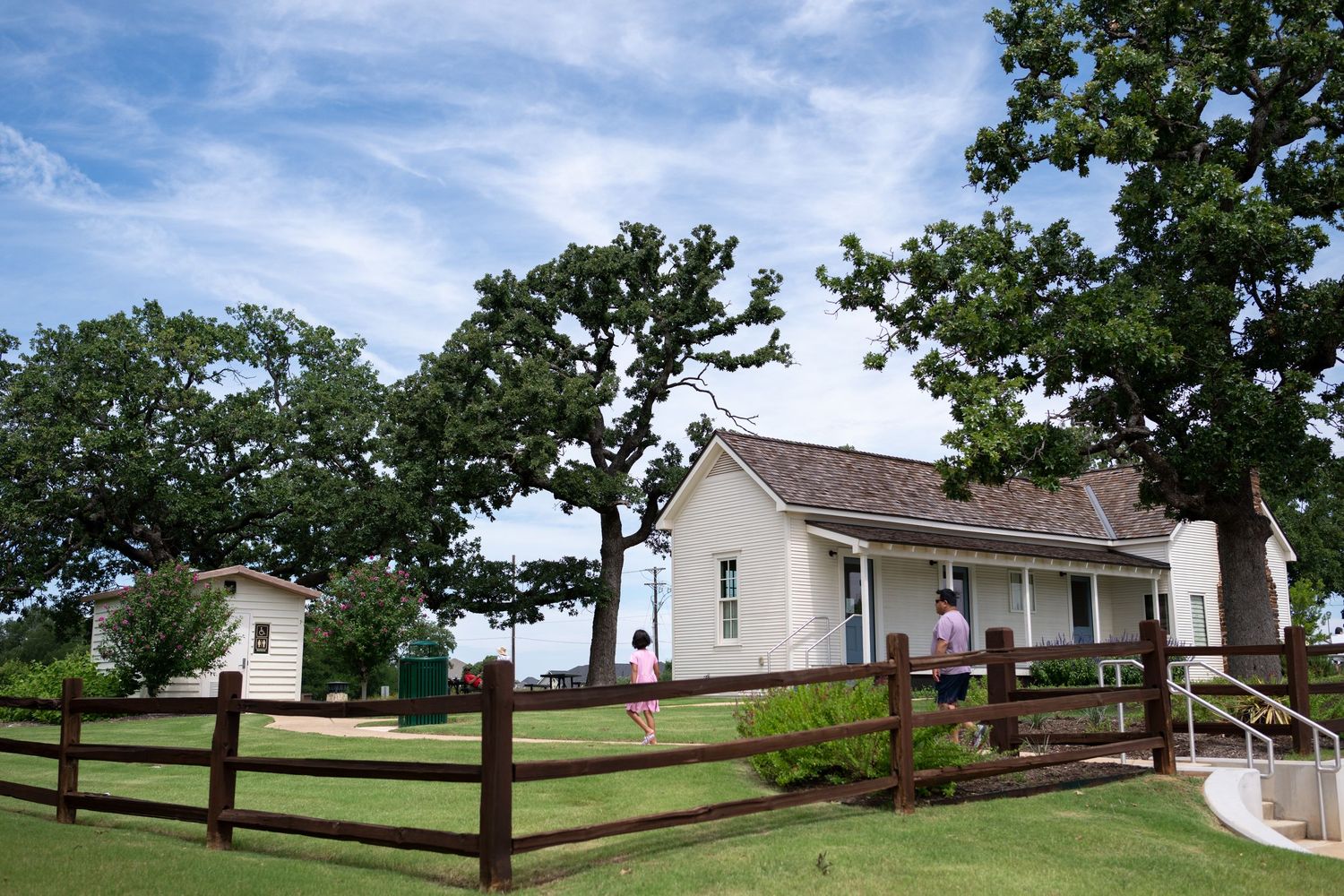 White farmhouse with wood fence, oak trees, and green lawn under blue sky.