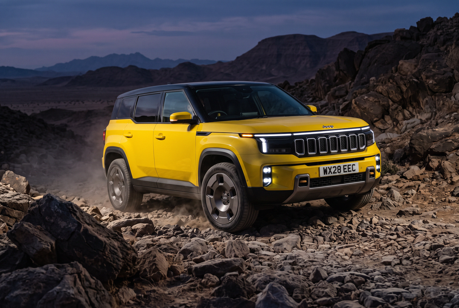 Yellow Jeep Grand Cherokee SUV navigating rocky desert terrain with mountain backdrop at dusk.