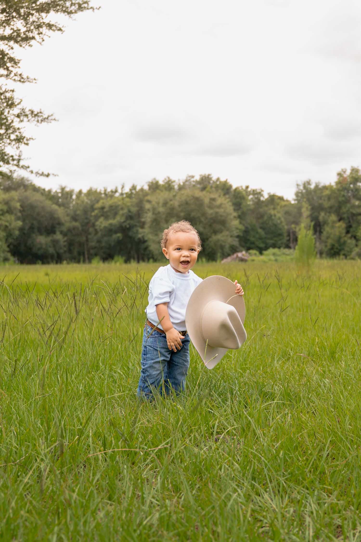 A happy toddler in jeans and white shirt holds a cowboy hat while standing in a lush green field.