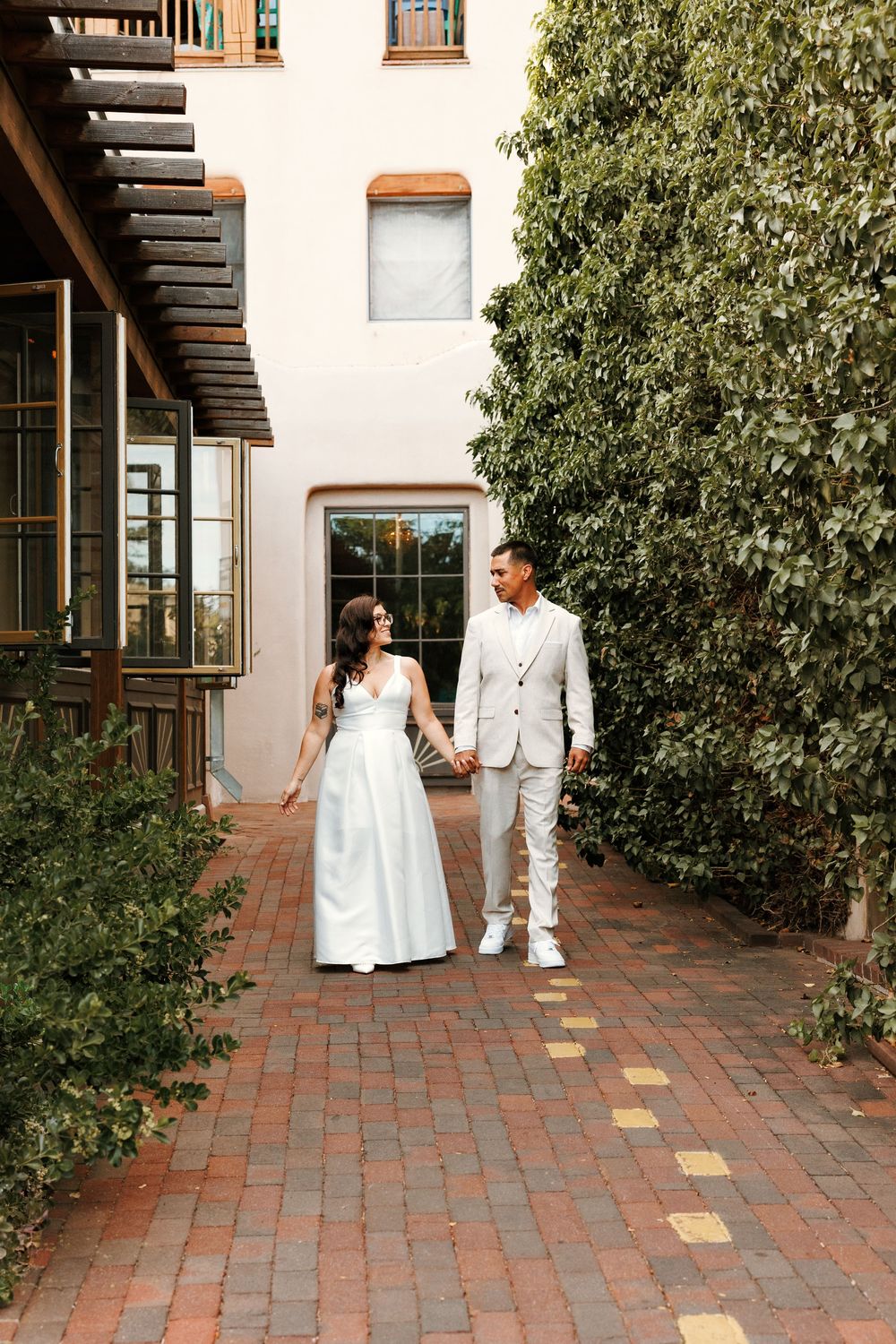 Newlyweds holding hands and walking along a brick courtyard path beside a building and tall hedges.
