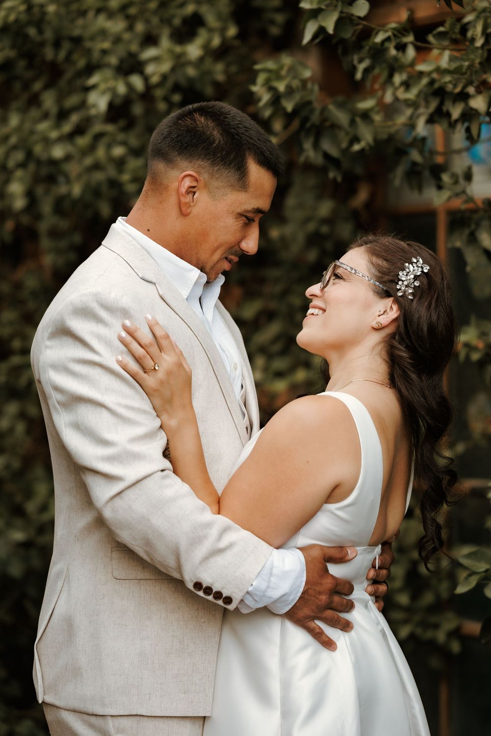Smiling bride in white dress with hair accessory embraces groom in beige suit outdoors.