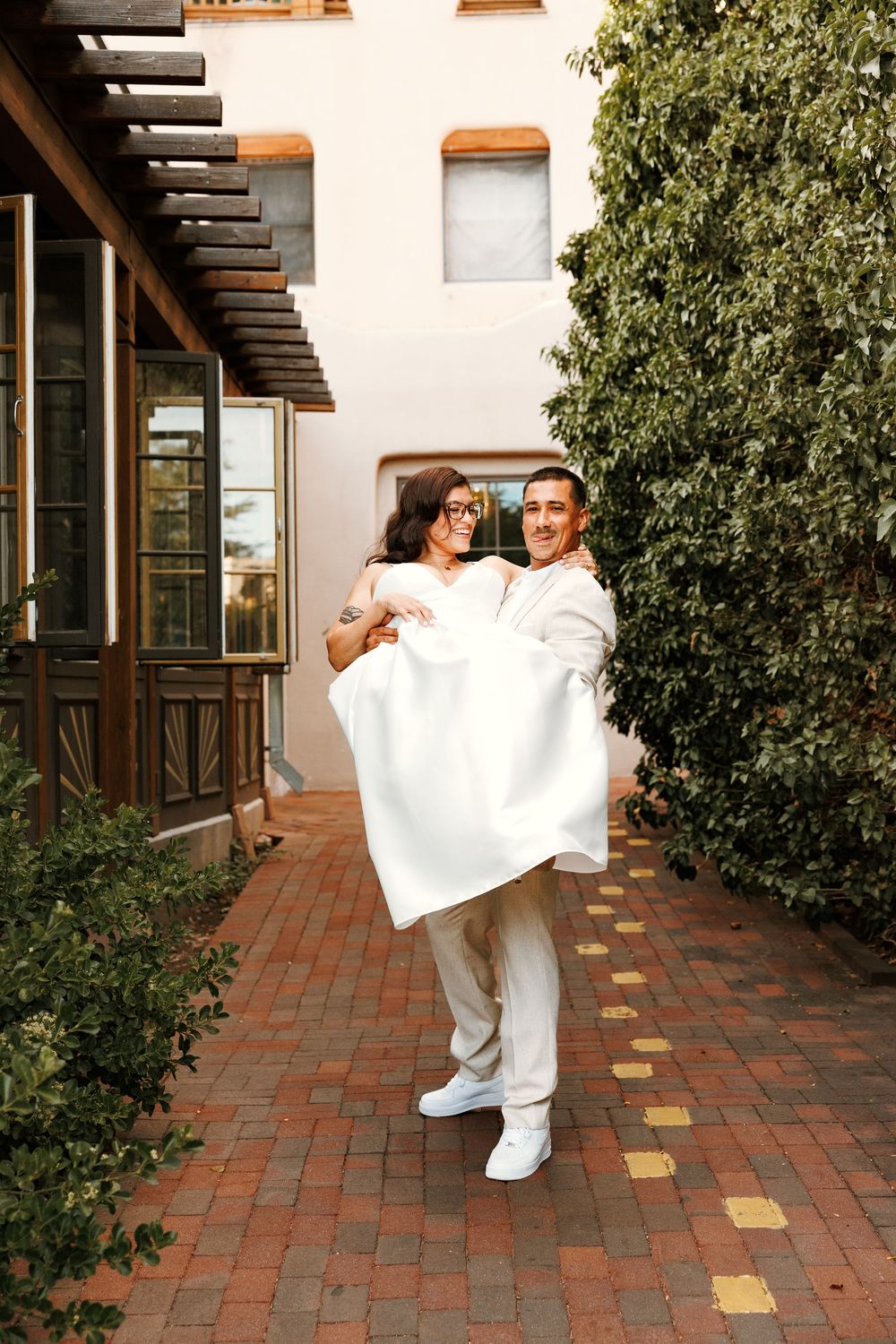 Groom carries bride in white dress outdoors on brick path near greenery and building on wedding day.