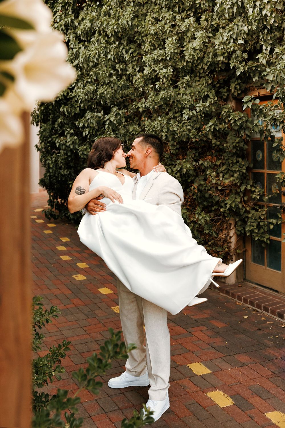 Groom in white suit lifts bride in wedding dress, sharing a romantic kiss outdoors against lush green ivy wall.