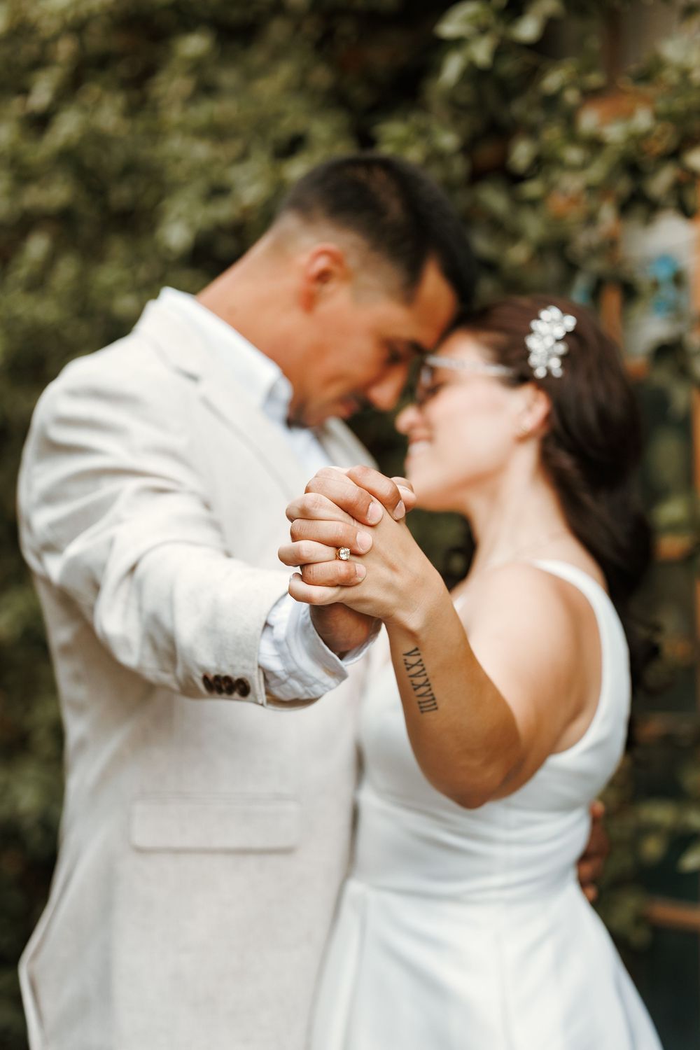 Bride and groom share first dance, holding hands with foreheads touching, outdoors with lush greenery backdrop.