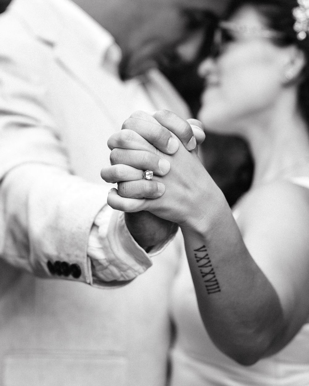 Black and white photo of newlyweds holding hands, showcasing engagement ring and Roman numeral tattoo during first dance.