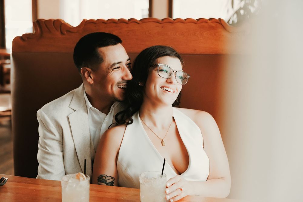 A happy couple sits together at a restaurant table, the man in a white blazer embracing the smiling woman wearing glasses.