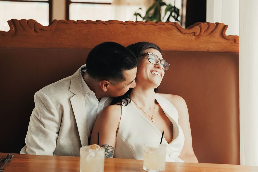 Groom kissing smiling bride on cheek at restaurant booth during romantic wedding day celebration.