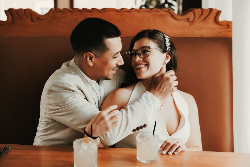 Smiling couple in wedding attire sharing a romantic moment at a restaurant booth with cocktails.