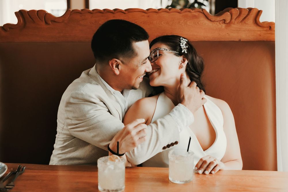 A joyful couple in wedding attire shares a romantic moment at a restaurant booth with cocktails on the table.