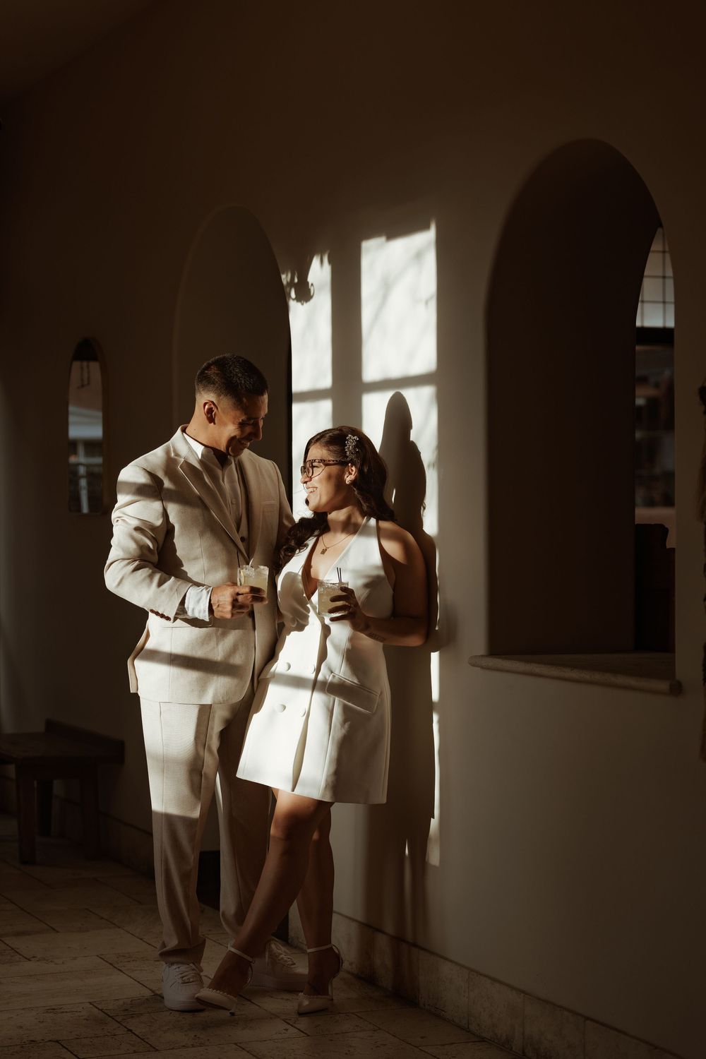 Elegant couple in white outfits sharing a romantic moment in a sunlit archway corridor.