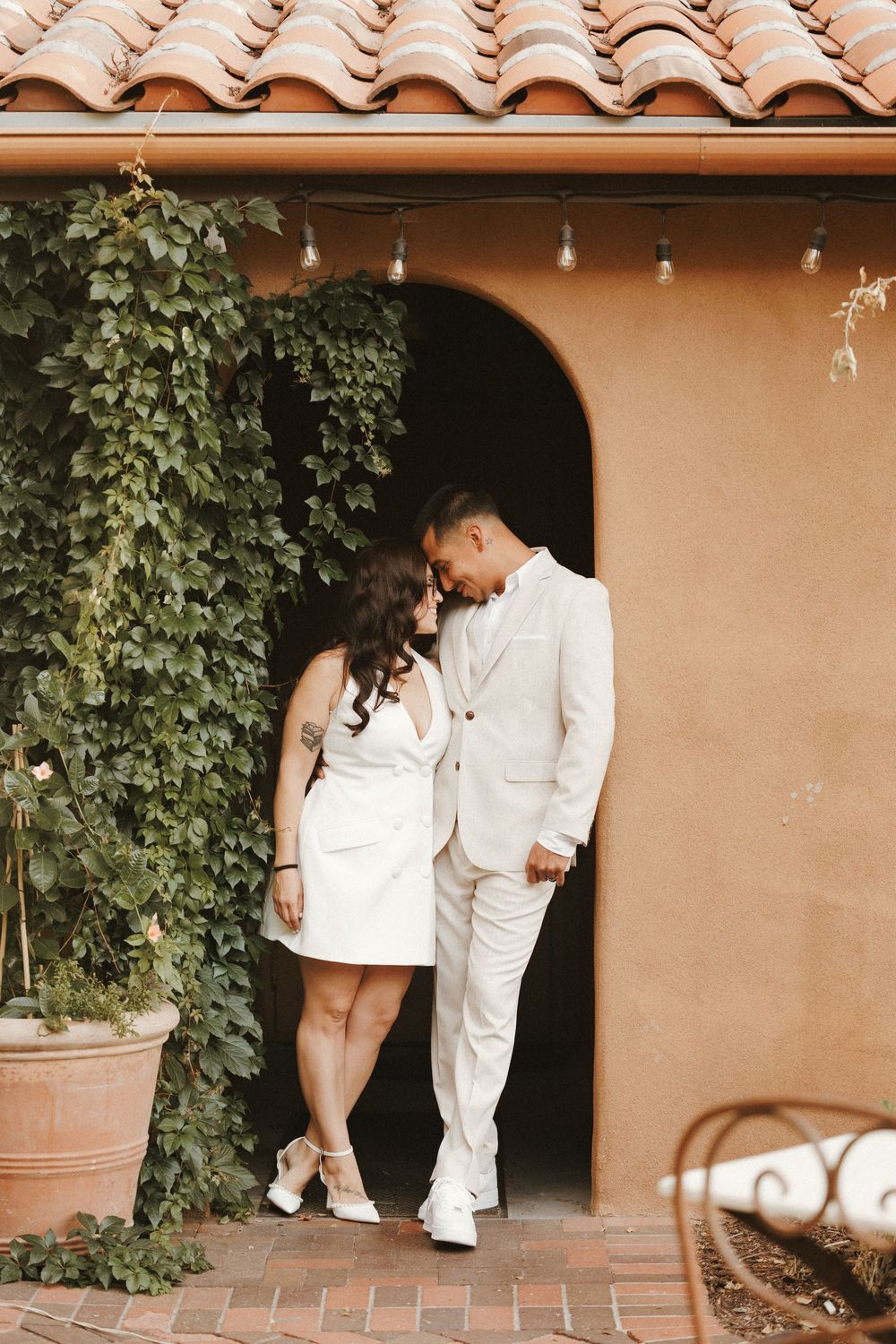 Bride and groom in white wedding attire standing close together in an arched doorway with ivy.