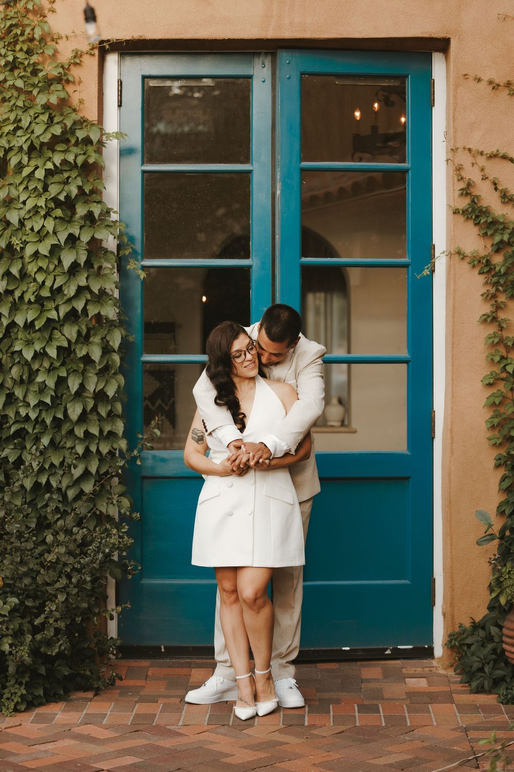 Couple embracing in front of a teal blue door surrounded by ivy on a warm beige wall, dressed in white outfits.