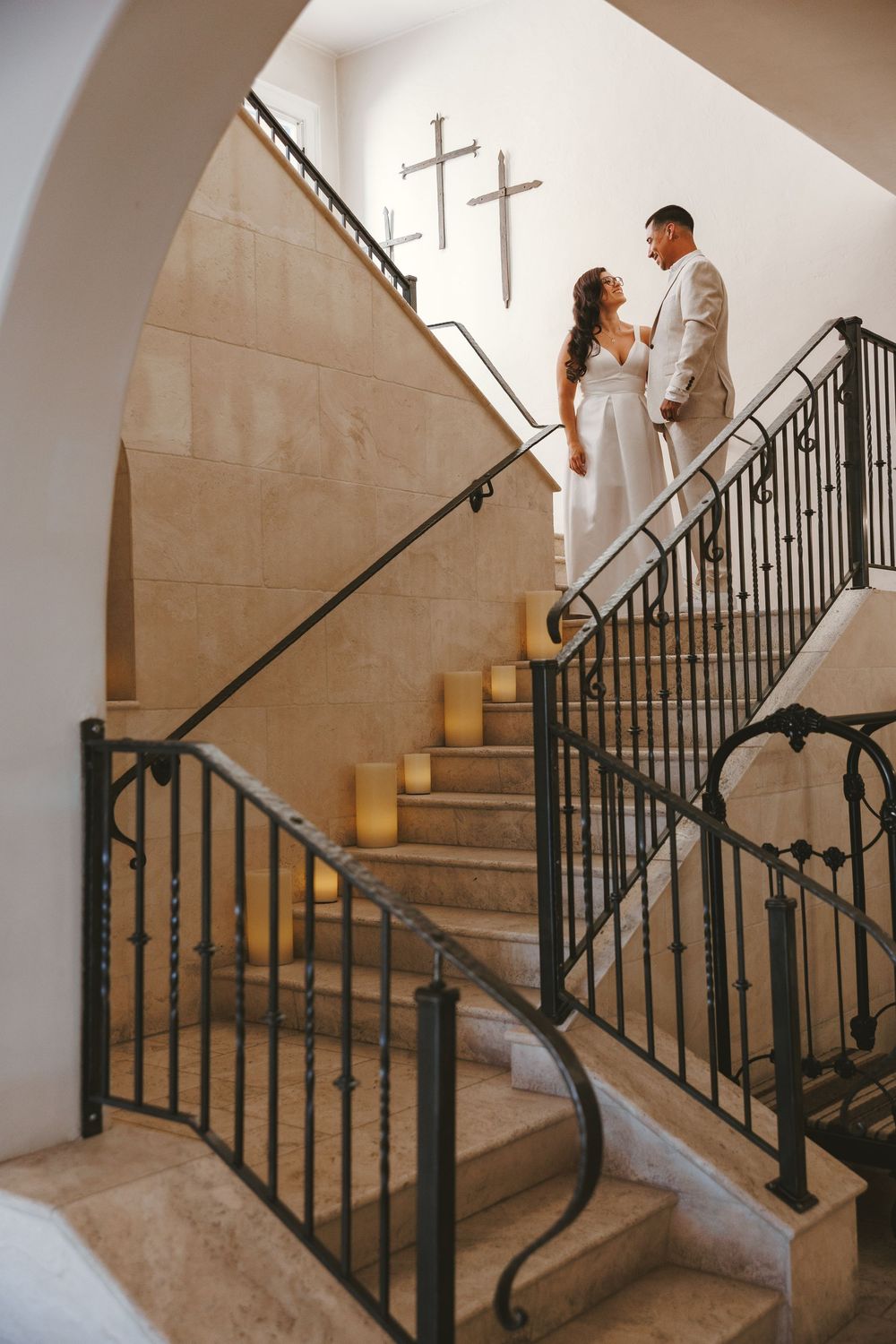 Bride and groom stand on candlelit stone staircase with iron railings, crosses on wall behind them.