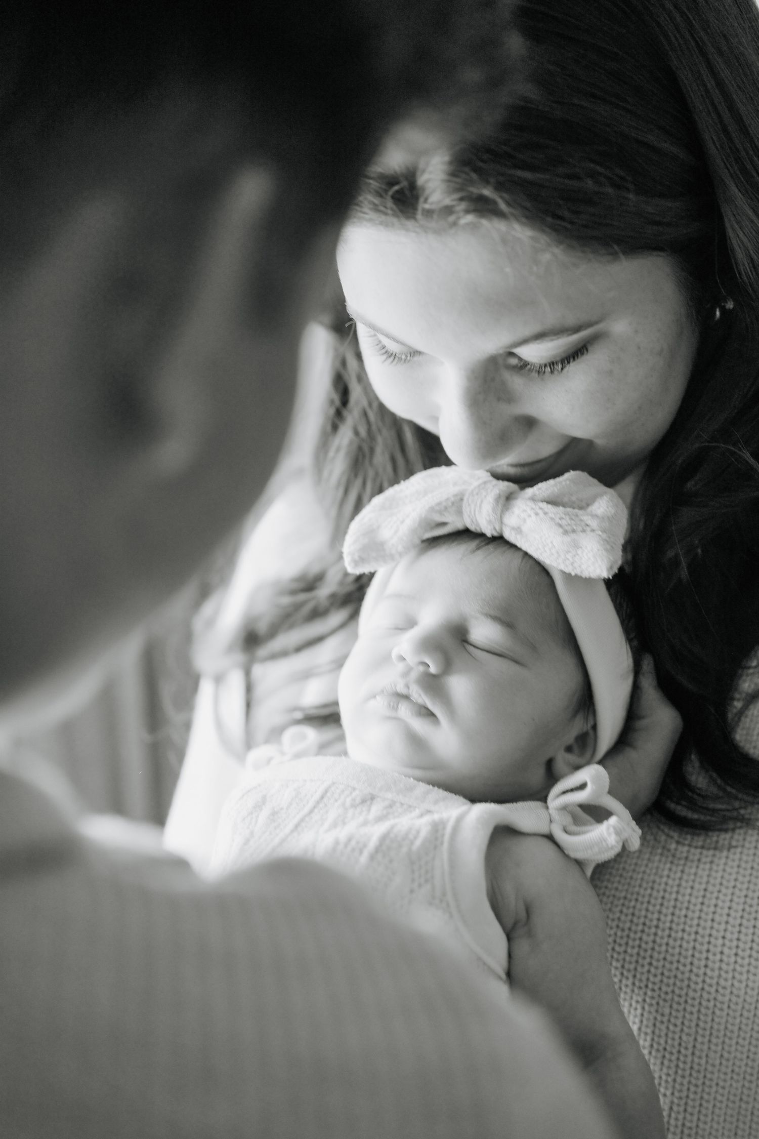 Black and white photo of a mother tenderly kissing her sleeping newborn baby wearing a bow headband.