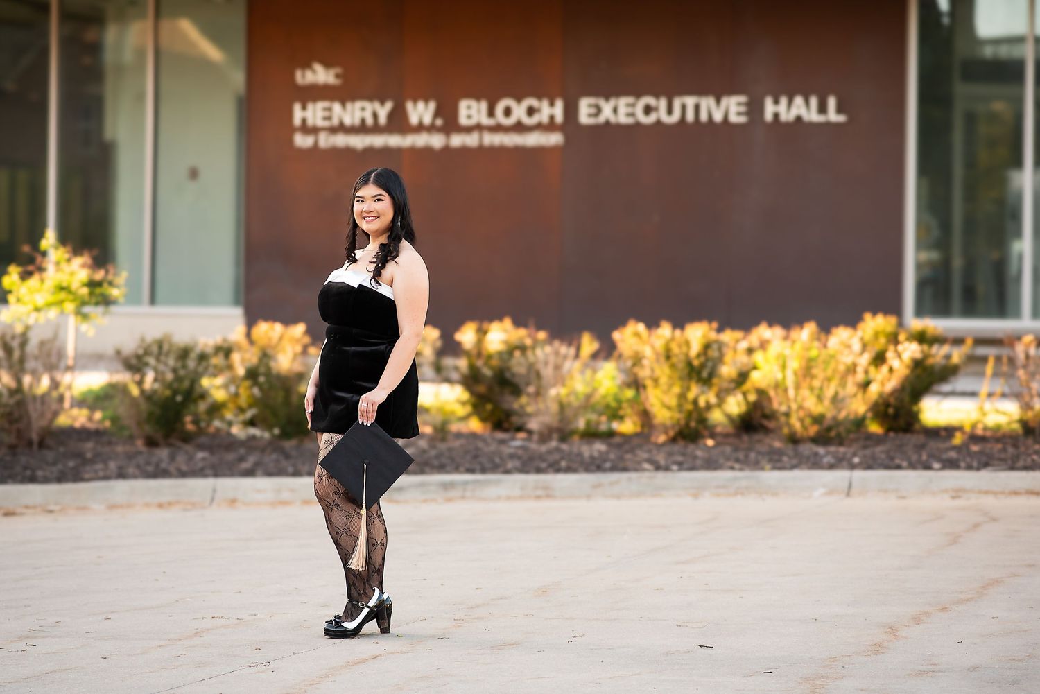 Woman in black dress posing outside Henry W. Bloch Executive Hall at UMKC on a sunny day.