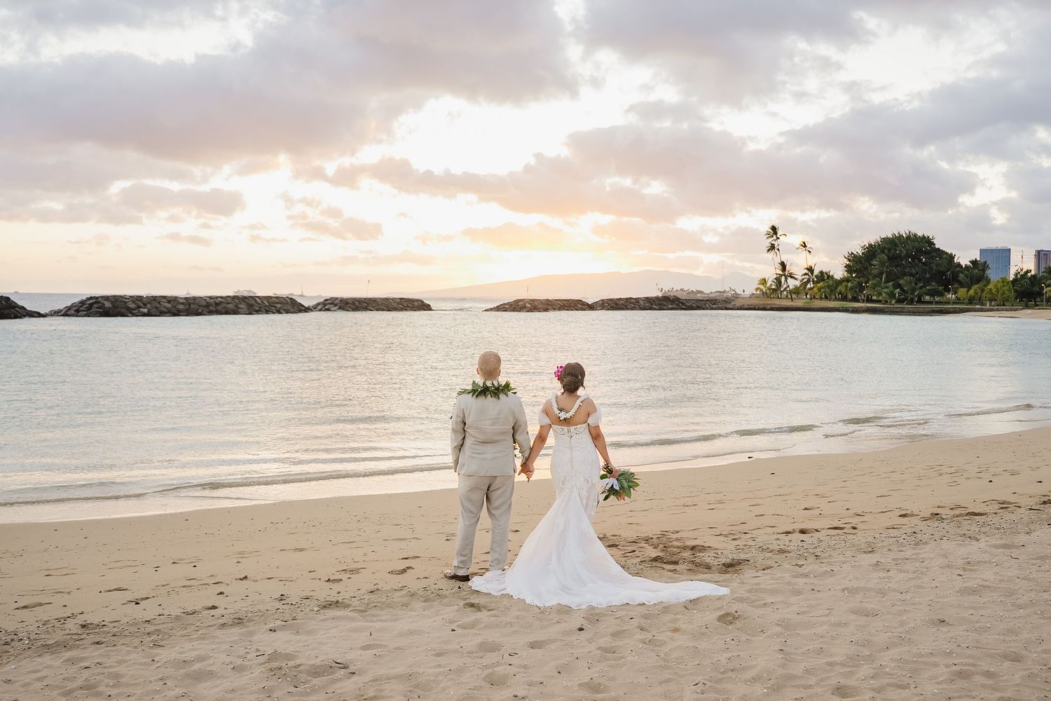 Bride and groom holding hands on a sandy Hawaiian beach at sunset in wedding attire.