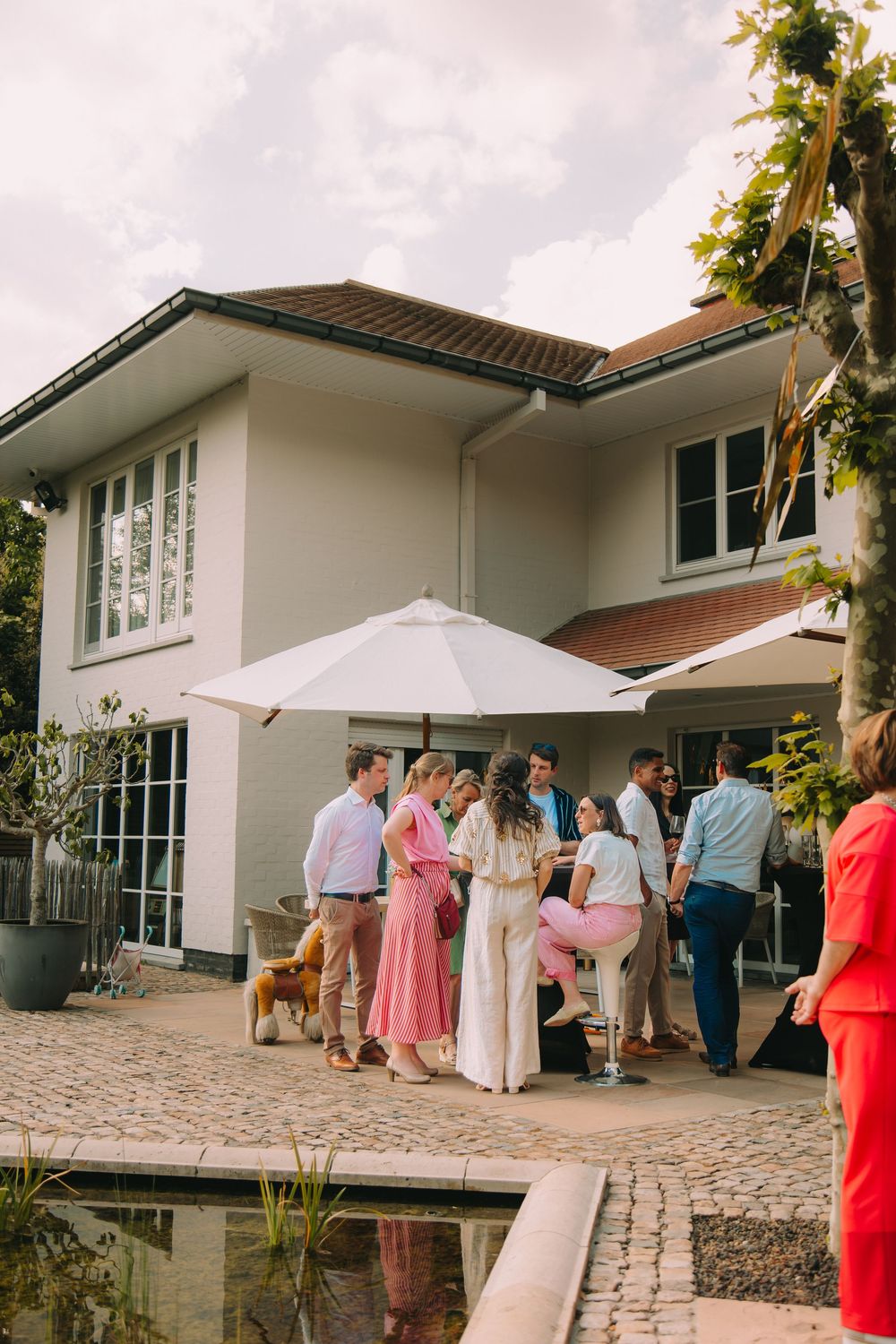Guests gather under a white patio umbrella at an elegant outdoor garden party beside a modern white house.