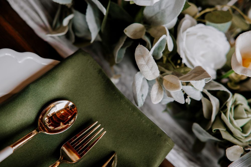 Elegant wedding table setting with green linen napkin, gold cutlery, and white floral arrangement.