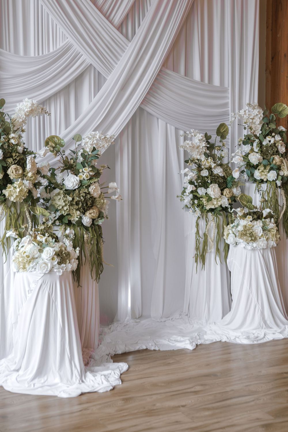 Elegant wedding arch with white draping and floral arrangements featuring roses and greenery on pedestals.