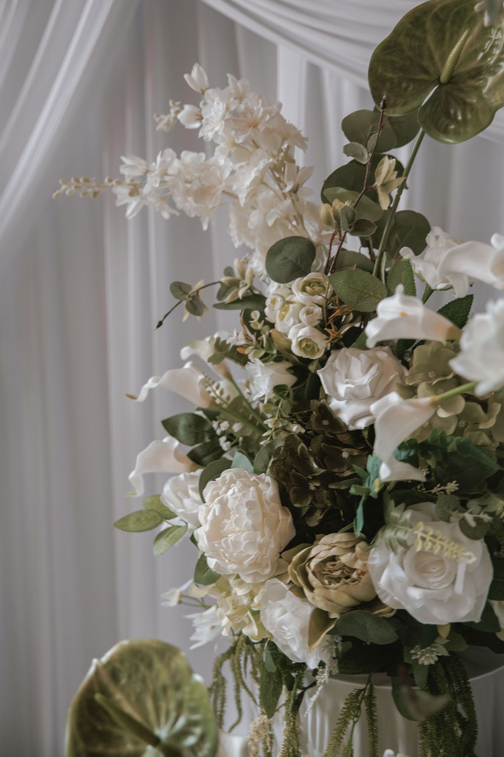 Elegant wedding floral arrangement featuring white peonies, roses, and greenery against a soft white draped backdrop.