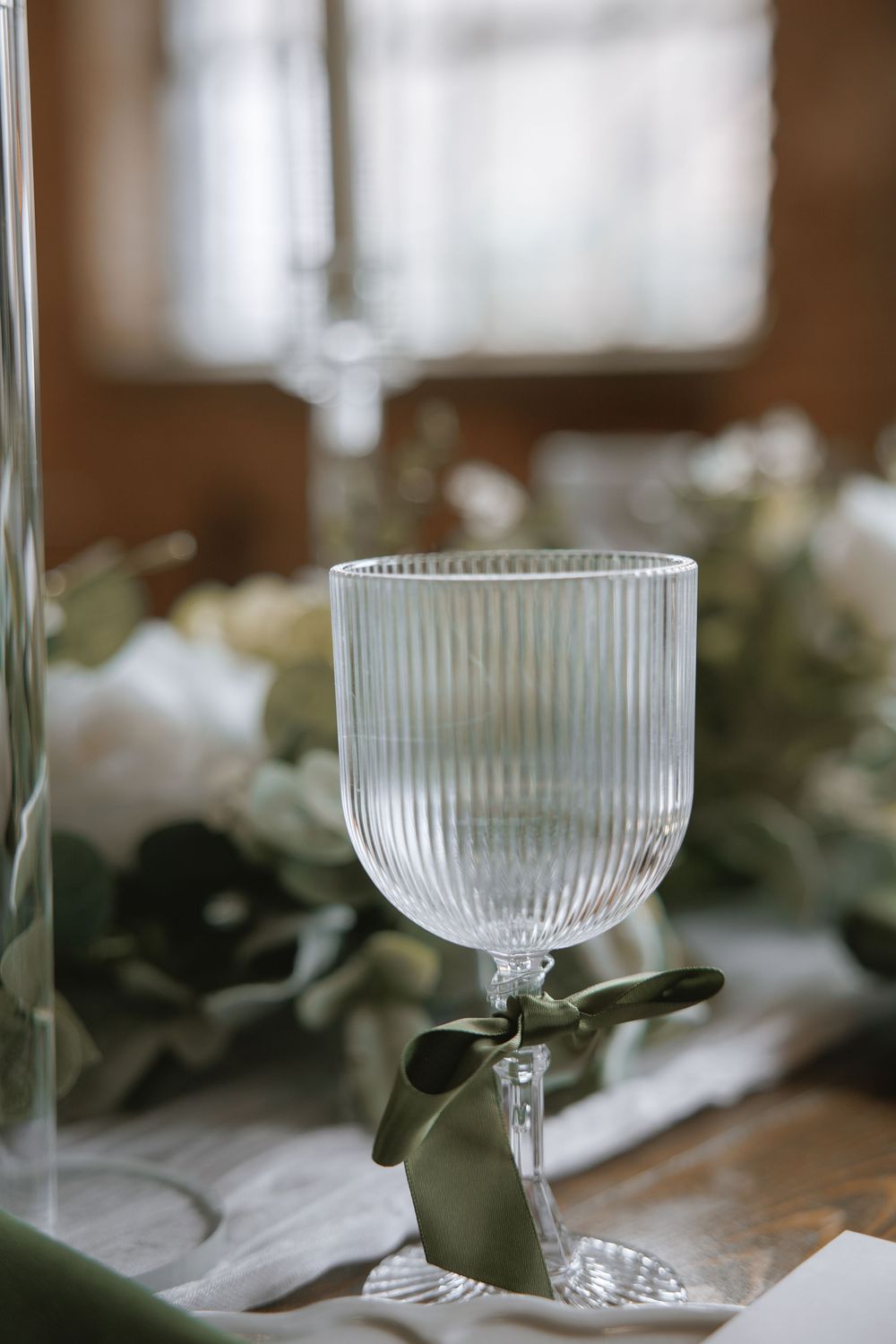 Elegant ribbed glass goblet with sage green ribbon bow on wedding table with white floral arrangement.