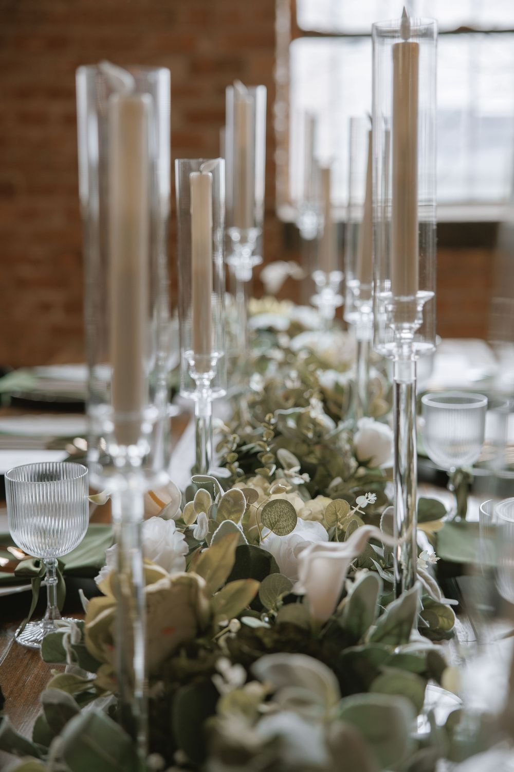Elegant wedding table with glass taper candle holders and lush greenery floral runner in a rustic brick venue.