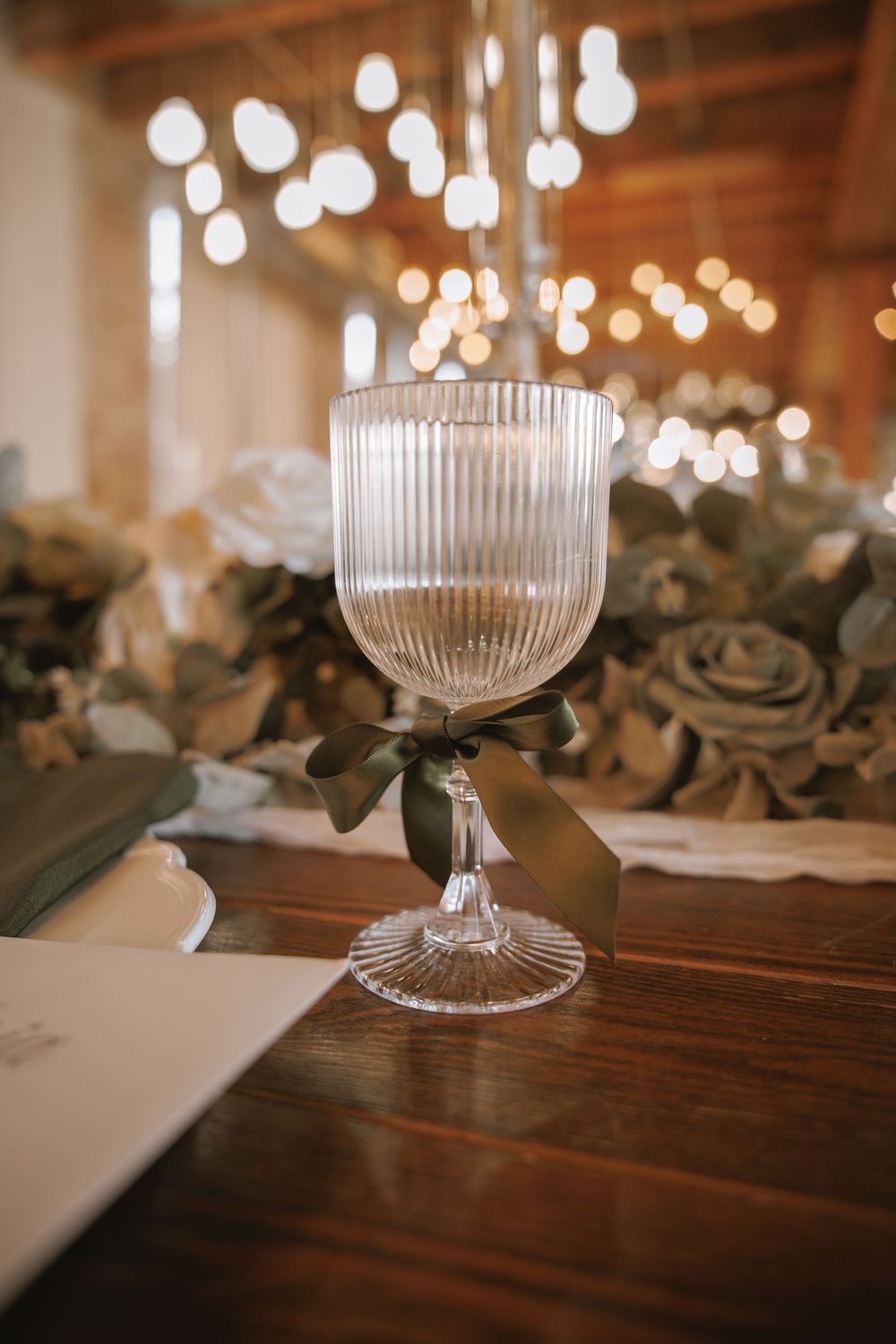 Ribbed crystal wine glass with green ribbon bow on wooden table with floral centerpiece and bokeh lights in background.