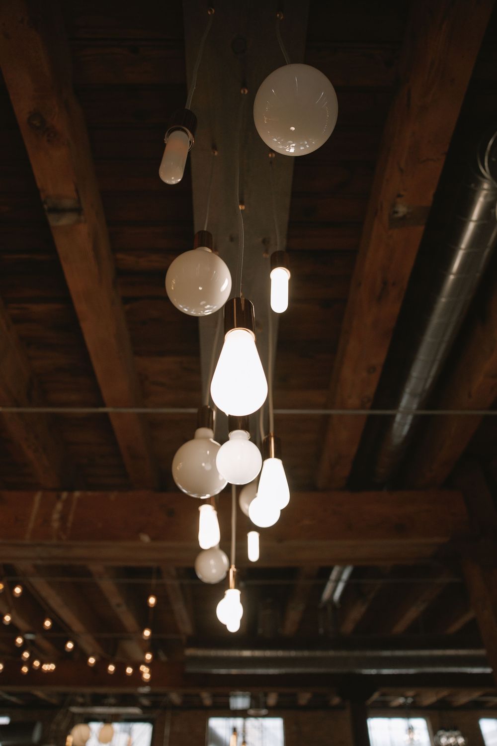 Modern pendant lights of varying shapes hang from rustic wooden beam ceiling in a warmly lit wedding venue.