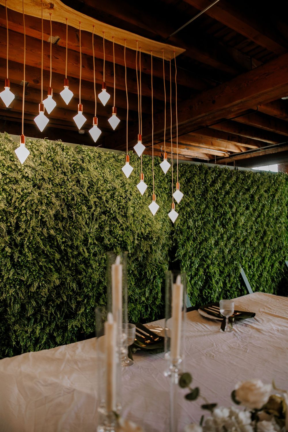 Wedding reception table with white candles beneath hanging geometric ornaments against a lush green moss wall.