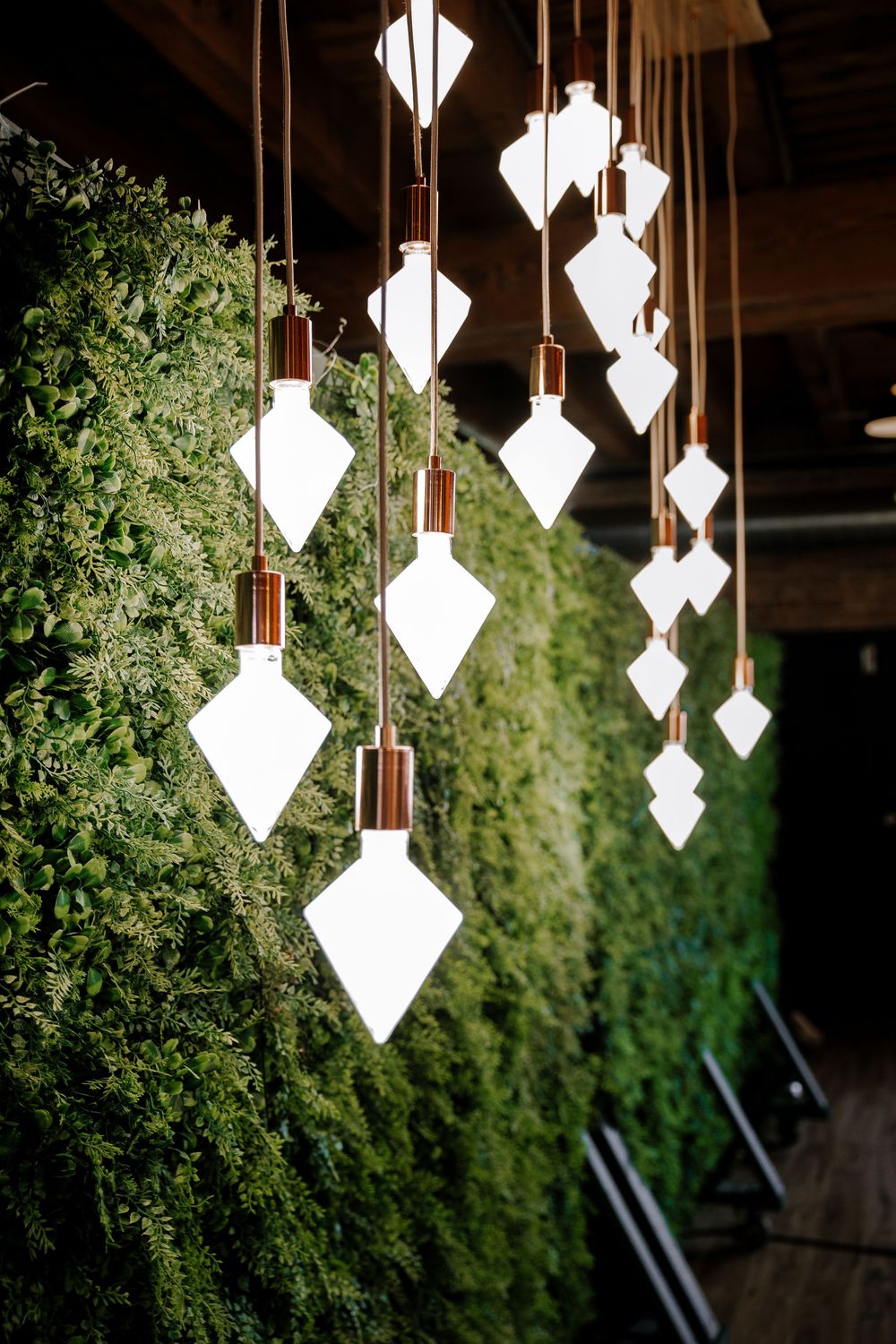 Diamond-shaped white pendant lights hang from copper fixtures against a lush green living plant wall backdrop.