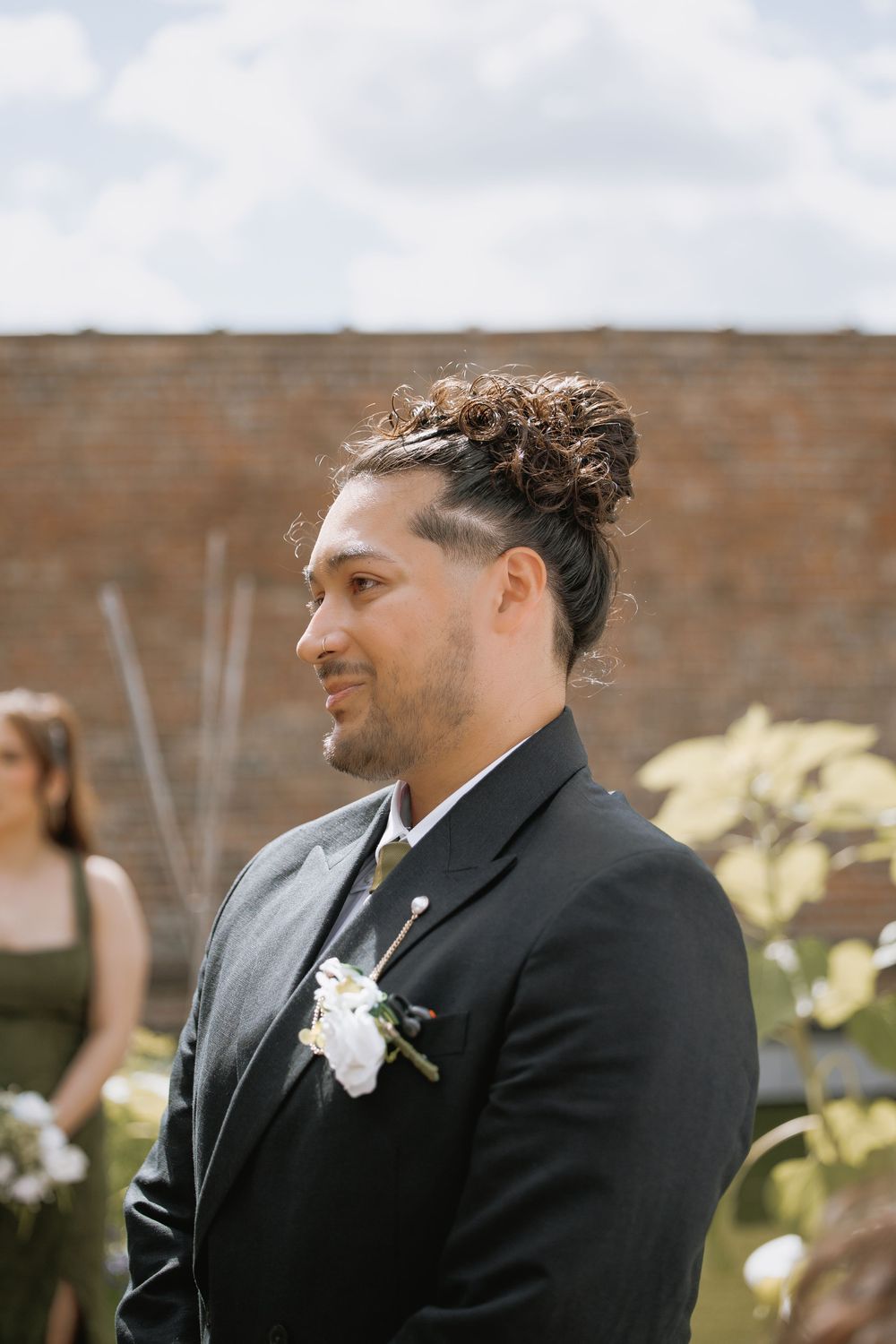 Groom in black suit with white boutonniere, hair in bun, standing outdoors at wedding ceremony near brick wall.