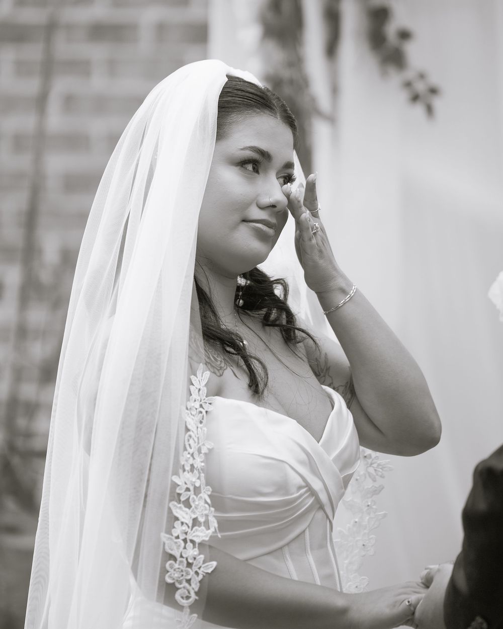 Emotional bride in white wedding dress and veil wiping tears during ceremony in black and white photo.