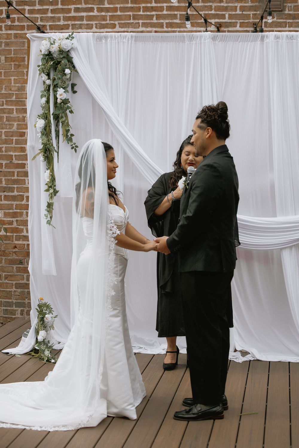 Bride and groom exchange vows before a white draped altar adorned with greenery and white flowers, officiated by a woman in black.