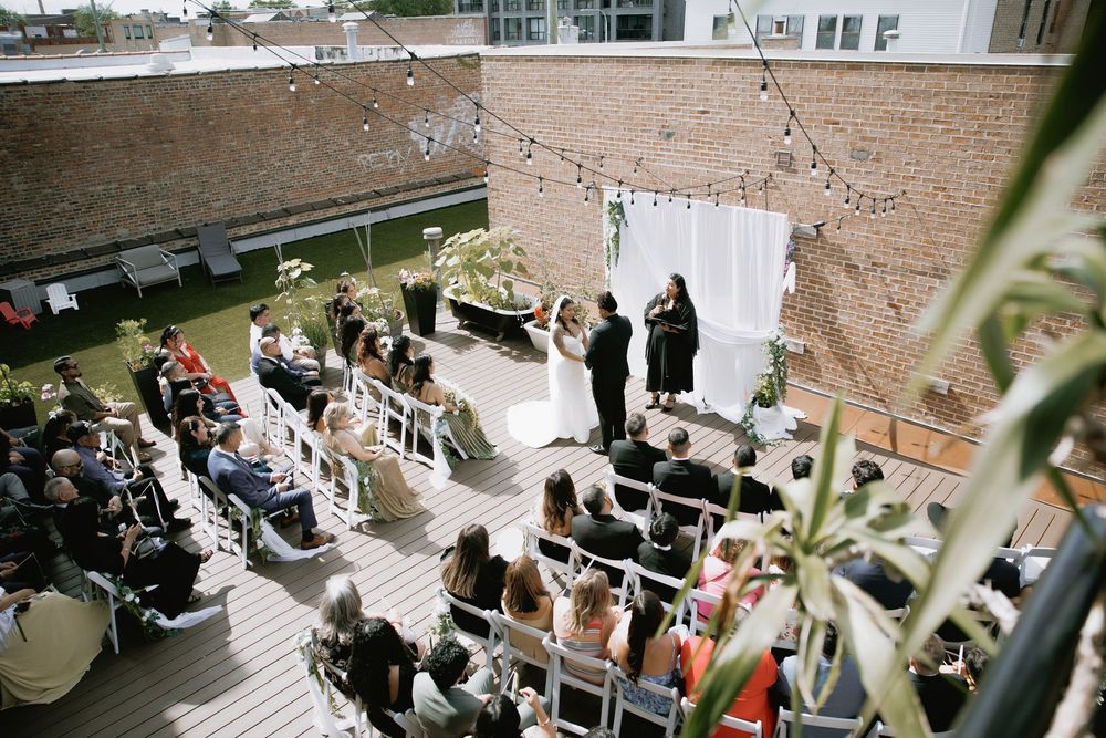 Outdoor rooftop wedding ceremony with guests seated in white chairs, couple at altar with white drape backdrop and string lights.