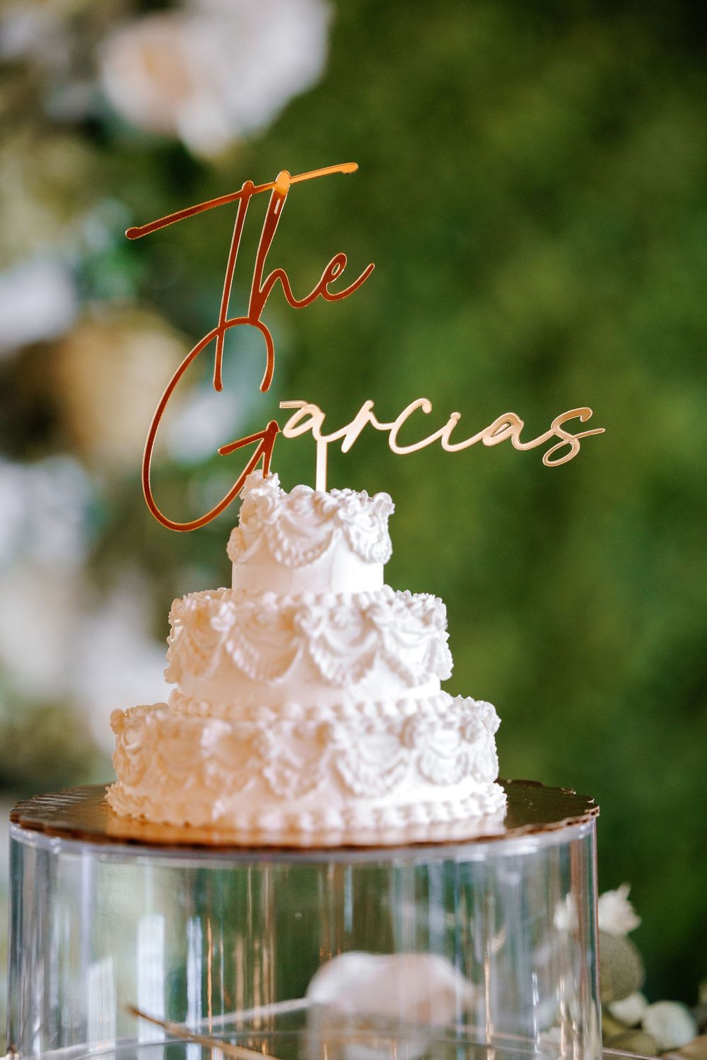 Elegant three-tier white wedding cake with ruffled frosting topped with gold "The Garcias" cake topper on glass stand.