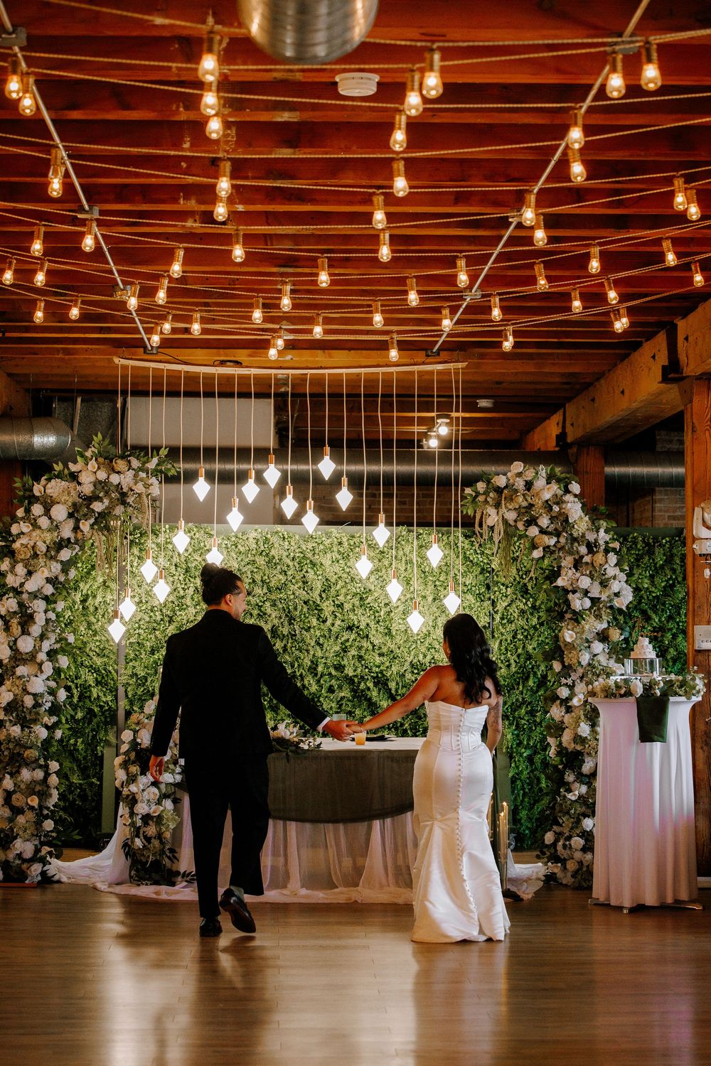 Bride and groom hold hands facing a floral arch with greenery wall, string lights, and hanging bulbs at wedding reception.