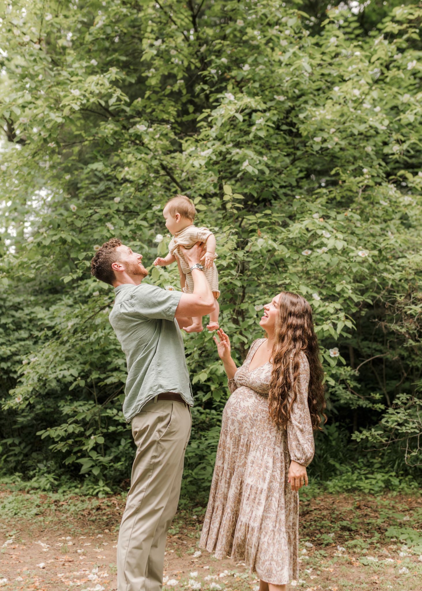 Pregnant woman and man lifting toddler in the air outdoors in a lush green forest setting.