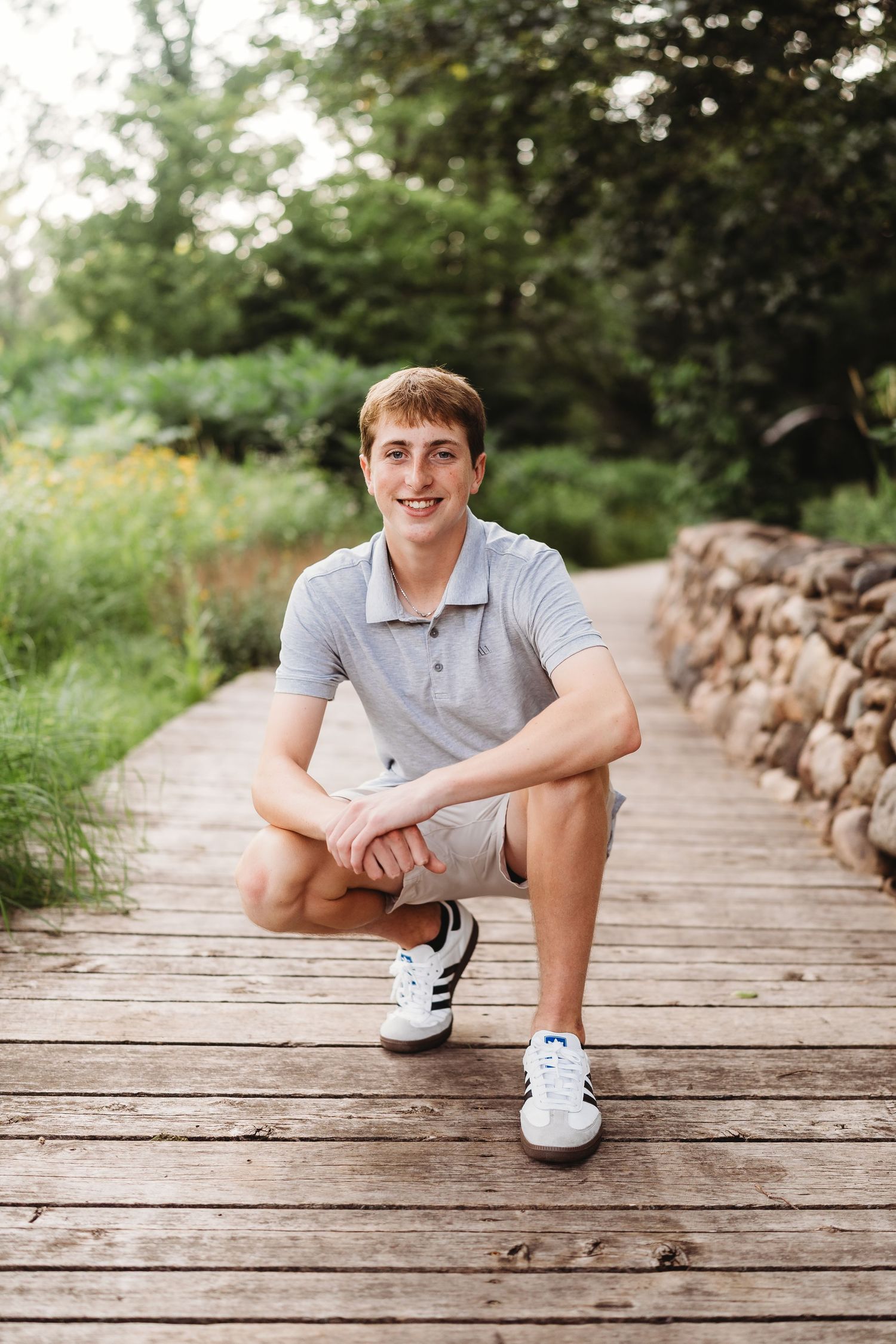 Young man in gray polo and shorts crouching on a wooden boardwalk path, smiling outdoors.