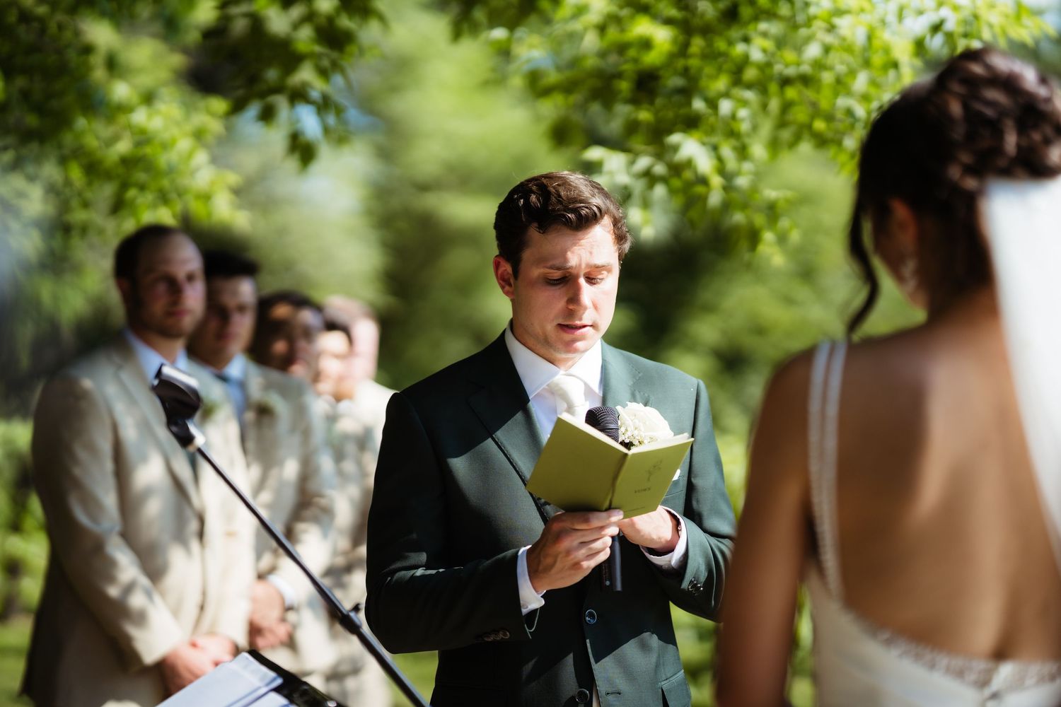 Groom in dark suit reads vows from a book during an outdoor wedding ceremony with guests seated behind.