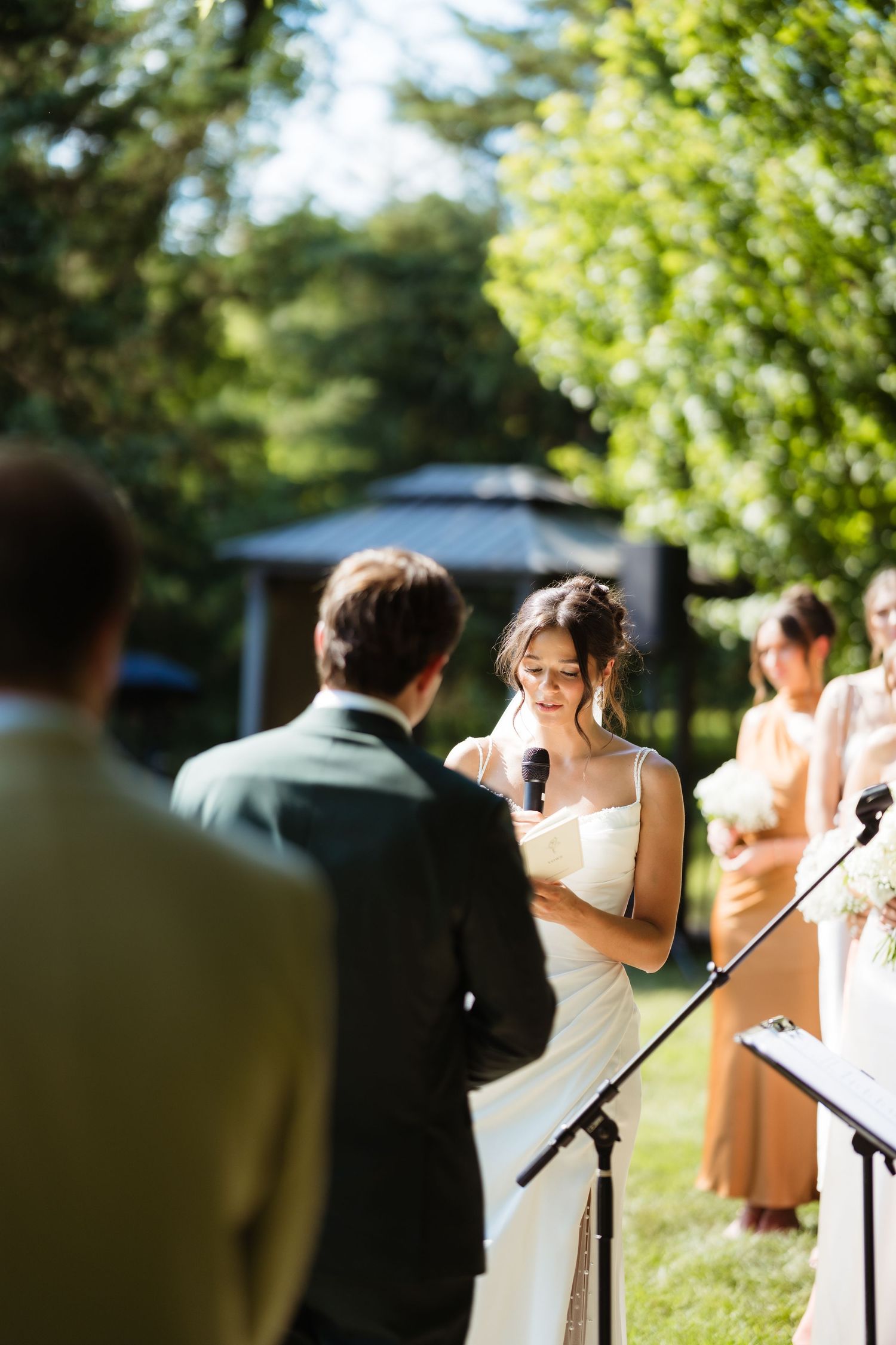 Emotional bride reading vows at outdoor wedding ceremony, groom in green suit, bridesmaids in gold dresses, lush garden backdrop.