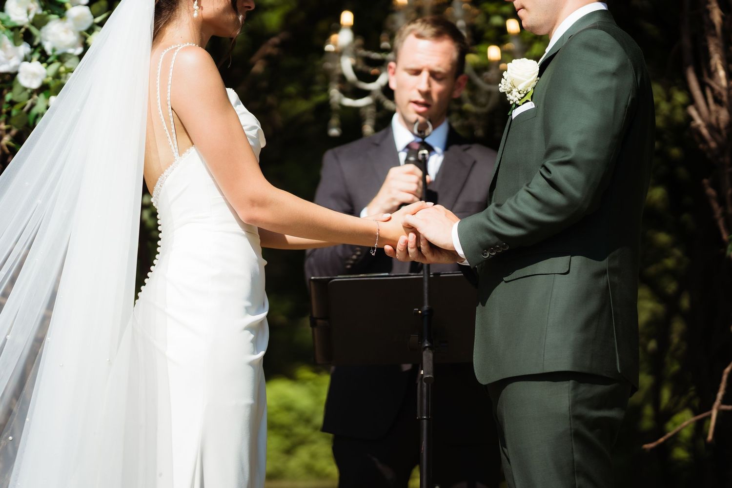 Bride and groom exchanging rings at outdoor wedding ceremony with officiant holding microphone.