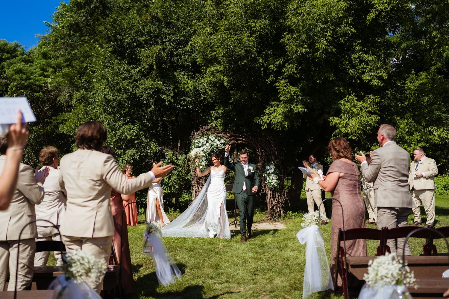 Outdoor wedding ceremony on green lawn with bride and groom walking the aisle surrounded by guests and lush trees.